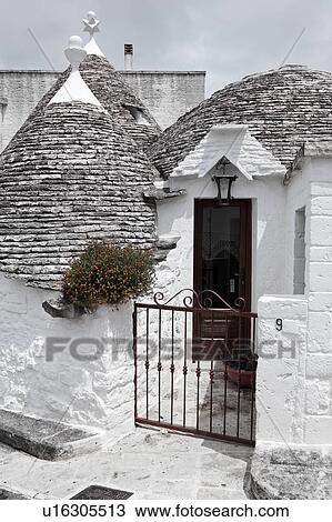 Stock Image - Street view: town of Alberobello, World Heritage site, with trullo house, white painted stone with piled stone conical roofs, wrought iron gate leading to front door, flowering green rockplant set against monochrome view, Puglia, Southern Italy. Fotosearch
