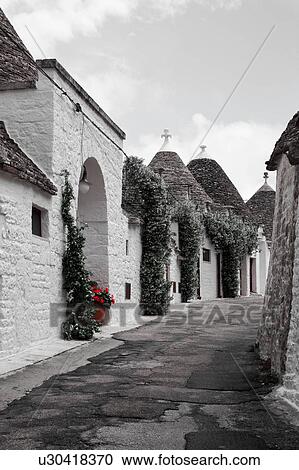 Stock Image - Street view: town of Alberobello, World Heritage site, with trullo houses, white painted stone with piled stone conical roofs, paved street and red flowers set against monochrome view, Puglia, Southern Italy. Fotosearch