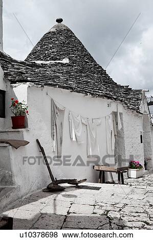 Stock Photo - Street view: town of Alberobello, World Heritage site, with trullo houses, white painted stone with piled stone conical roofs, paved street, laundry hanging and farm utensils against wall, red flowers set against monochrome view, Puglia, Southern Italy. Fotosearch