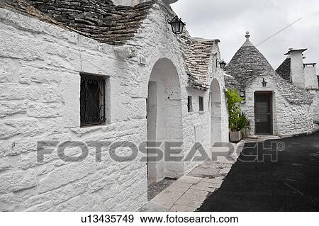 Stock Photo - Street view: town of Alberobello, World Heritage site, with trullo houses, white painted stone with piled stone conical roofs and large chimneys, paved and asphalt street, green plants set against monochrome view, Puglia, Southern Italy. Fotosearch