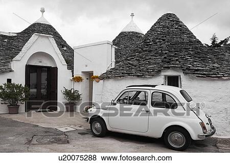 Stock Photo - Street view: town of Alberobello, World Heritage site, with trullo houses, white painted stone with piled stone conical roofs, paved street, yellow flowers, white traditional car set against monochrome view, Puglia, Southern Italy. Fotosearch