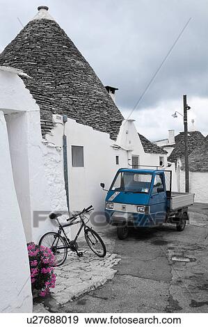 Street view: town of Alberobello, World Heritage site, with trullo houses, white painted stone with piled stone conical roofs, paved street and red flowers, bicycle and blue traditional van set against monochrome view, Puglia, Southern Italy View Large Photo Image Stock Photo - Street view: town of Alberobello, World Heritage site, with trullo houses, white painted stone with piled stone conical roofs, paved street and red flowers, bicycle and blue traditional van set against monochrome view, Puglia, Southern Italy. Fotosearch