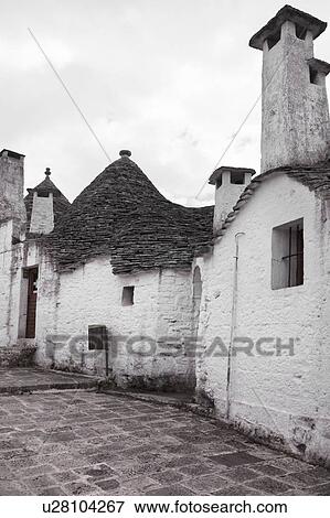 Stock Photo - Street view: town of Alberobello, World Heritage site, with trullo houses, white painted stone with piled stone conical roofs and large chimneys, paved street, monochrome view, Puglia, Southern Italy. Fotosearch