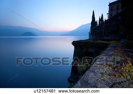 Stock Image - Tranquil scene overlooking Lake Como at sunset, blue and pink sky, still waters, cypress trees, wall with trailing ivy with autumn foliage, Lake Como, Northern Italy. Fotosearch