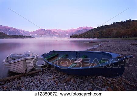 Stock Image - Two traditional wooden fishing boats beached at shore of lake in pink dawn glow, with mountain range and opposite shoreline reflected in calm waters of Lake Como, Northern Italy. Fotosearch