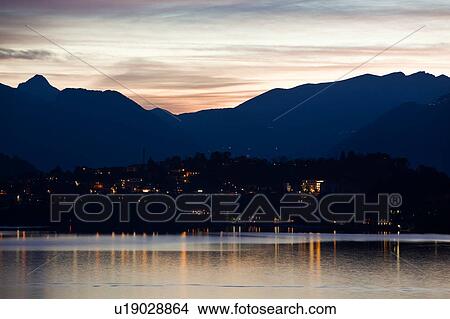 Picture - View across Lake Como at dusk, with beautiful calm sunset colours of blue, purple and pastel pink, mountains silhouetted beyond Bellagio, with town lights illuminated, Lake Como, Northern Italy. Fotosearch