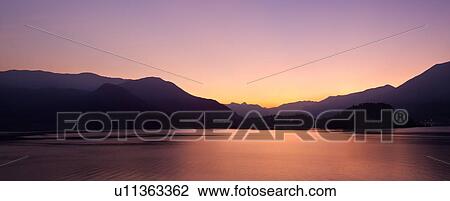Stock Image - View across Lake Como at dusk, with beautiful calm sunset colours of purple and pastel pink, mountains silhouetted beyond Bellagio, with town lights illuminated, Lake Como, Northern Italy. Fotosearch