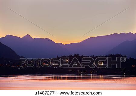 Stock Image - View across Lake Como at dusk, with beautiful calm sunset colours of purple and pastel pink, mountains silhouetted beyond Bellagio, with town lights illuminated, Lake Como, Northern Italy. Fotosearch