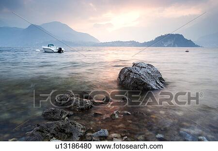 View across Lake Como at sunset, with beautiful pastel blue and pink sky, fishing boat in water, beyond rocky shore, mountains silhouetted beyond Bellagio, Lake Como, Northern Italy View Large Photo Image Stock Image - View across Lake Como at sunset, with beautiful pastel blue and pink sky, fishing boat in water, beyond rocky shore, mountains silhouetted beyond Bellagio, Lake Como, Northern Italy. Fotosearch