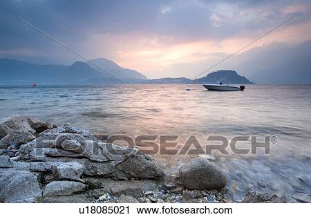 View across Lake Como at sunset, with beautiful pastel blue and pink sky, fishing boat in water, beyond rocky shore, mountains silhouetted beyond Bellagio, Lake Como, Northern Italy View Large Photo Image Stock Image - View across Lake Como at sunset, with beautiful pastel blue and pink sky, fishing boat in water, beyond rocky shore, mountains silhouetted beyond Bellagio, Lake Como, Northern Italy. Fotosearch