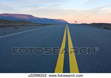 Stock Image - View along straight road from El Chalten at sunset, with view of mountain range, and blue sky with delicate pink cloud formation, Argentina, Patagonia. Fotosearch