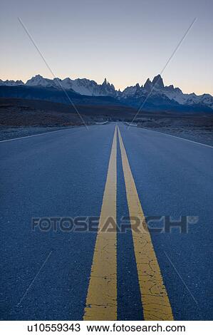 Stock Image - View along straight road to El Chalten at sunset, with view of Andes mountain range, with Mount Fitzroy and Cerro Torre backlit by setting sun, Argentina, Patagonia. Fotosearch