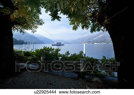 Picture - View of Bellagio lakefront in early morning sunshine, autumn colours of beech trees, passenger ferry leaving dock, mountain range visible across Lake Como, Northern Italy. Fotosearch
