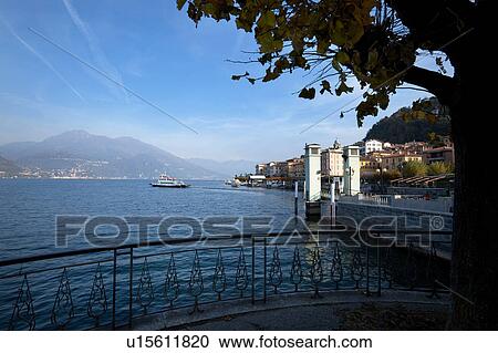Stock Image - View of Bellagio lakefront in early morning sunshine, autumn colours of beech trees, passenger ferry leaving dock, mountain range visible across Lake Como, Northern Italy. Fotosearch