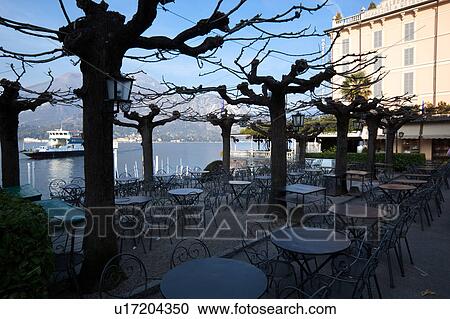 Stock Image - View of Bellagio lakefront seen from interior of outdoor cafe in early morning sunshine, autumn colours of beech trees, passenger ferry leaving dock, mountain range visible across Lake Como, Northern Italy. Fotosearch