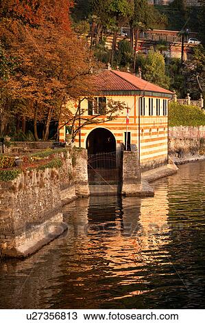 Stock Image - View of boat house of Villa Cipressi overlooking Lake Como on sunny afternoon, horsechestnut trees with autumn foliage, Lake Como, Northern Italy. Fotosearch