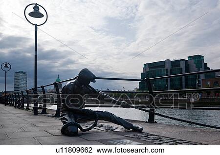 Stock Image - View of Campshire Walk, Statue of The Linesman, with wrought iron railings and street lamps at sunset with River Liffey, modern buildings beyond, overcast dramatic sky, Dublin, Southern Ireland. Fotosearch