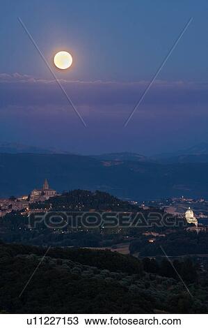 Stock Image - View of full moon rising over medieval hilltop town of Todi, with spire of San Fortunato, and Tempio of Bramante, town lights illuminated at dusk, Umbria, Italy. Fotosearch