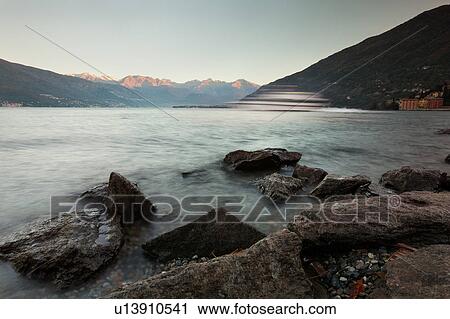 View of lake from rocky shoreline, wave and ferry boat motion slowed, pink light of sunrise on mountain range across Lake Como, Northern Italy View Large Photo Image Stock Image - View of lake from rocky shoreline, wave and ferry boat motion slowed, pink light of sunrise on mountain range across Lake Como, Northern Italy. Fotosearch