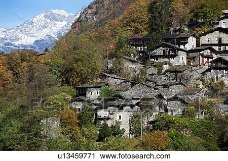Stock Image - View of mountain village with stone houses, slate roofs, nestled amongst trees with autumnal foliage, glacier beyond on mountain slope on autumn sunny morning, Lombardy, Northern Italy. Fotosearch