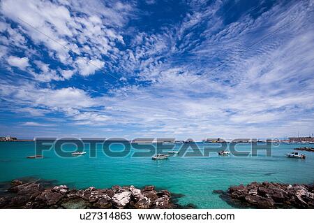 Stock Image - View of Otranto harbour with clear green water, moored sailboats, cargo boat, and motorboats, sunny spring morning with dramatic clouds in blue sky, Salento, Puglia, Southern Italy. Fotosearch