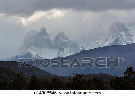 Stock Photograph - View of snow covered Los Cuernos mountains with threatening autumn sky, late afternoon, with hills and trees silhouetted in foreground, Torres del Paine, Southerm Chile, Patagonia. Fotosearch