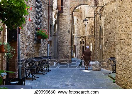 Stock Image - View of stone paved street with medieval stone buildings lining each side, and connecting arch above, with wrought iron lamp and window grille, decorative red flowers, backview of lady carrying shopping, Todi, Umbria, Italy. Fotosearch