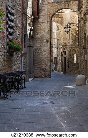Stock Photo - View of stone paved street with medieval stone buildings lining each side, and connecting arch above, with wrought iron lamp and window grille, decorative red flowers, Todi, Umbria, Italy. Fotosearch