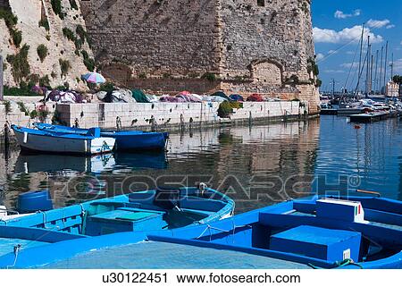 Stock Image - Wooden fishing boats moored in harbour, with fishing nets on quayside below the medieval walls of Gallipoli, and sailboats beyond, sunny spring morning, Salento, Puglia, Southern Italy. Fotosearch