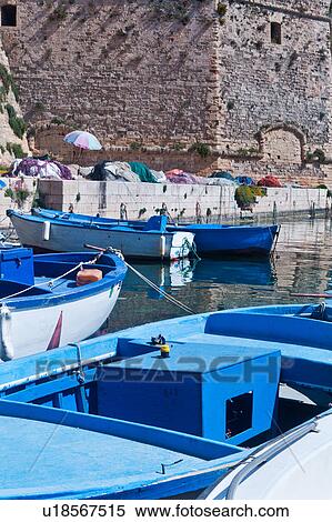 Stock Photography - Wooden fishing boats moored in harbour, with fishing nets on quayside below the medieval walls of Gallipoli, sunny spring morning, Salento, Puglia, Southern Italy. Fotosearch