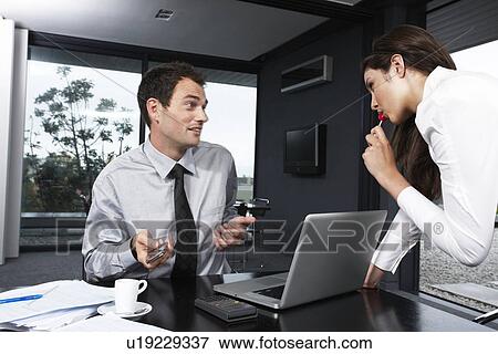 Stock Photo - Young businesswoman sucking on lollipop while leaning over young businessman's desk. Fotosearch