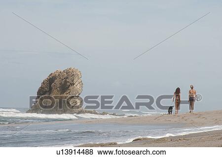 2 distant figures in swim suits strolling on the sandy beach on an overcast day with a leashed dog passing a rock stack View Large Photo Image Stock Photo - 2 distant figures in swim suits strolling on the sandy beach on an overcast day with a leashed dog passing a rock stack. Fotosearch