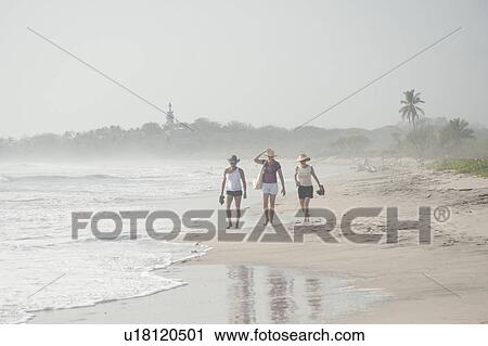 Stock Image - 3 women in shorts and hats walking on the beach. Fotosearch