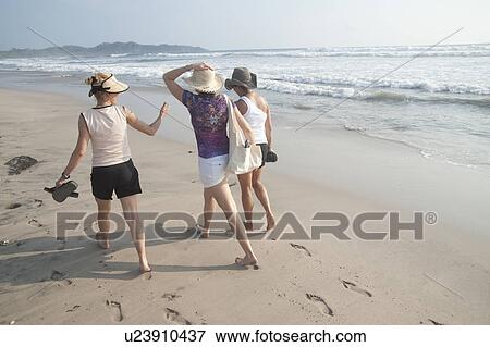Stock Photo - 3 women in shorts and hats walking on the beach. Fotosearch