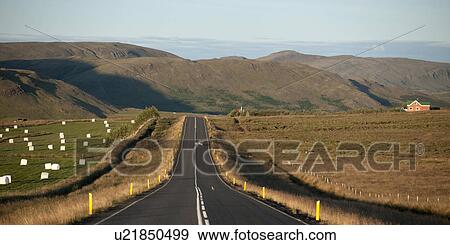 Stock Photo - Asphalt highway through farmland and hay bales, leading to mountains. Fotosearch