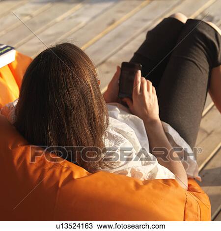 Stock Image - Back view of a woman resting on a dock looking at her cell phone at Lake of the Woods, Ontario. Fotosearch
