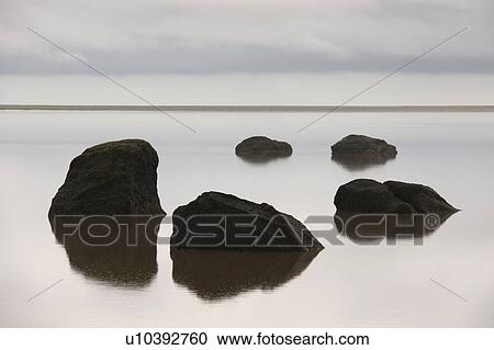 Stock Image - Circle of five black rocks rising from the ocean, mirrored in the water, horizon in the background. Fotosearch