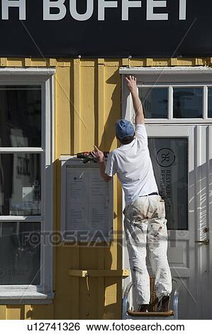 Stock Photograph - Commercial painter preparing door frame outside on a chair on a yellow building. Fotosearch