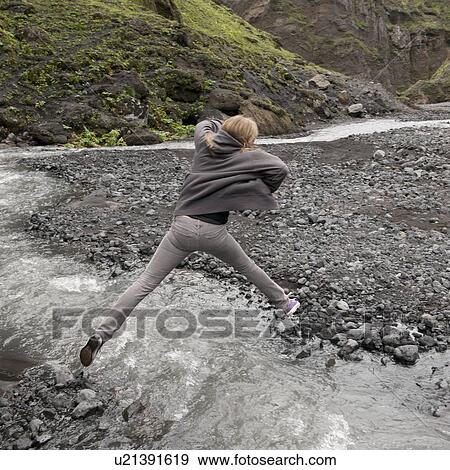Stock Photo - Girl leaping over fast flowing stream with rocky riverbank. Fotosearch
