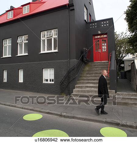 Stock Image - Icelandic businessman on sidewalk, past a black dwelling with red doors, and a wheelchair lift for the stairs, past the crosswalk with greens dots. Fotosearch