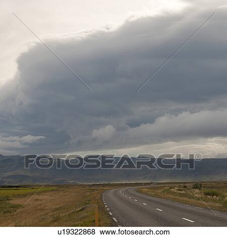 Stock Photo - Landscape, cloudy sky over highway on farmland driving towards mountains. Fotosearch