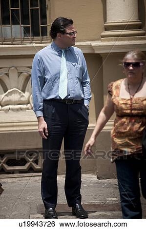 Stock Photograph - Man with a tie and woman in glasses on the sidewalk, a stone carved icanthus leaf on a building behind them, and an iron window grill. Fotosearch