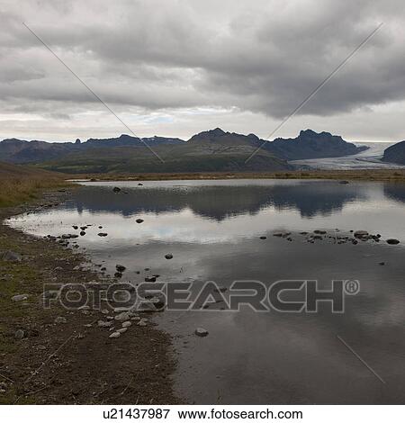 Stock Photo - Rocky shoreline of glacial lake, mountains and glacier in distance. Fotosearch
