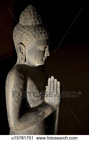Stock Image - Stone Buddha statue with hands in prayer position with strong shadows. Fotosearch