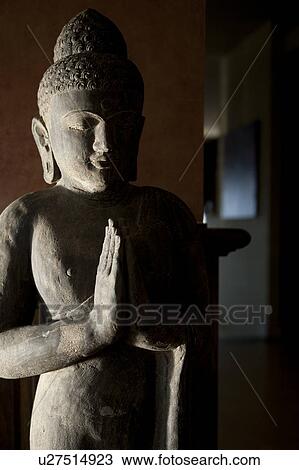 Stock Image - Stone Buddha statue with hands in prayer position with strong shadows. Fotosearch
