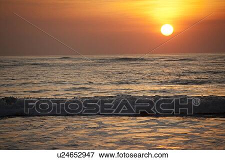 Stock Photo - Sun setting over the calm ocean with an orange glow. Fotosearch