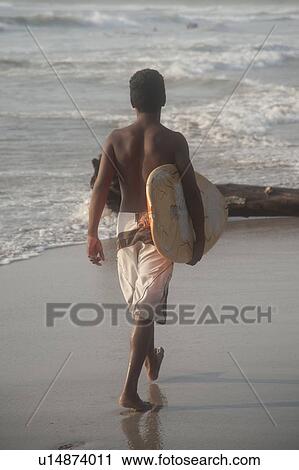 Stock Image - Teenage boy in a swim suit walking along the beach carrying a surfboard. Fotosearch