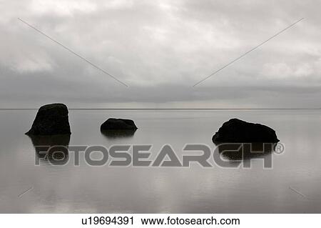 Stock Image - Three solitary rocks rising from the calm ocean against the horizon on a cloudy day. Fotosearch