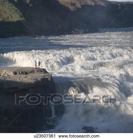 Stock Image - Whitewater flowing into waterfall in valley past rock outcrop with sightseers. Fotosearch