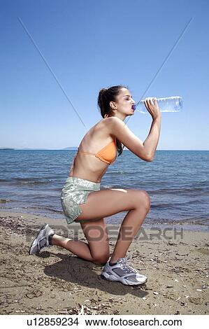 Picture - Athletic female young adult drinking bottled water on beach. Fotosearch